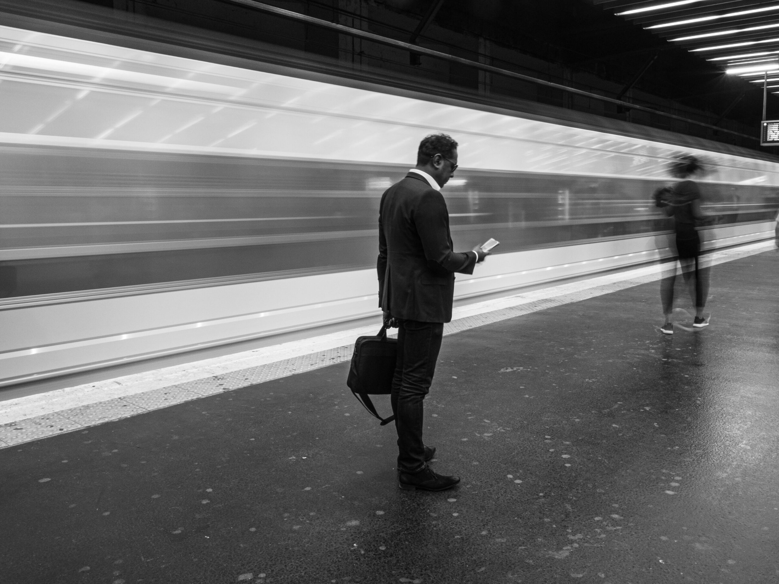 Black and white photo of a businessman at a bustling Paris metro station, holding his phone.
